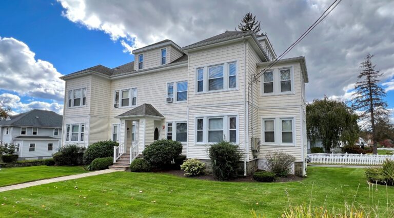 Large white multi-story house with a lawn and shrubbery.