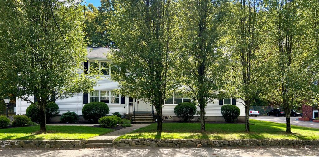 Two trees stand in front of a white house with a porch and green bushes.